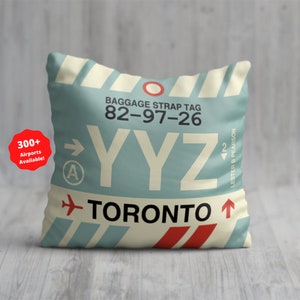 Peut inclure: Coussin carré avec un fond bleu clair, imitant une étiquette de bagage. Il affiche le texte "BAGGAGE STRAP TAG 82-97-26", "YYZ" et "TORONTO". Le design comprend un cercle rouge et un avion. Un autocollant indique "300+ Airports Available!"