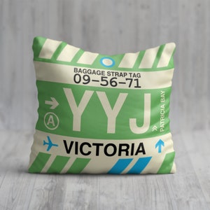 May include: A beige pillow with a green and white striped design. The pillow has a luggage tag design with the text "BAGGAGE STRAP TAG 09-56-71 YYJ VICTORIA PATRICIA BAY" and an airplane icon.