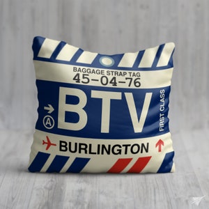 May include: A cream-coloured pillow with a blue and red luggage tag design. The tag has the text "BTV" in large white letters, "BURLINGTON" in smaller white letters, and the text "BAGGAGE STRAP TAG 45-04-76" in black letters. The tag also has a blue circle with a white arrow pointing to the right and the letter "A" inside the circle. The tag has the text "FIRST CLASS" in black letters on the right side.