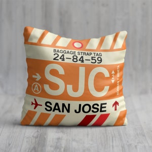 May include: Orange and white cushion with a luggage tag design. The tag has the airport code "SJC" and the text "San Jose" with an aeroplane icon. The tag also has the text "Baggage Strap Tag 24-84-59" and "Norman Y. Mineta".