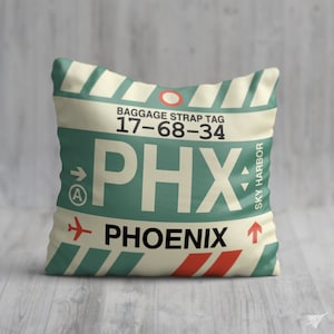 May include: A white cushion with a green and red luggage tag design. The tag says "Baggage Strap Tag 17-68-34 PHX SKY HARBOR PHOENIX".