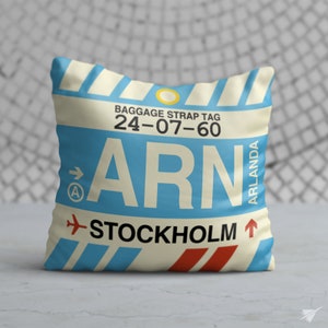 May include: A white pillow with a blue and red luggage tag design. The tag says "ARN" and "STOCKHOLM" with the airport code "24-07-60" and "ARLANDA" printed on the tag.
