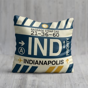 May include: A white pillow with a blue and yellow luggage tag design. The tag says "BAGGAGE STRAP TAG 21-36-60 IND INDIANAPOLIS" with an airplane icon and an arrow pointing up. The tag also has a priority label with an arrow pointing up.