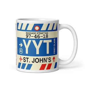 May include: White ceramic mug with a baggage strap tag design. The mug features the text "Yyt St. John's" in blue, red, and white, along with the numbers "97-46-18" and the words "First Class".