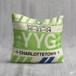 May include: A white pillow with a green luggage tag that says "YYG CHARLOTTETOWN" and "BAGGAGE STRAP TAG 36-21-04" in black text. The tag also has a blue arrow pointing to the right and a blue circle with a white letter "A" inside. The tag also has the words "FIRST CLASS" in black text.