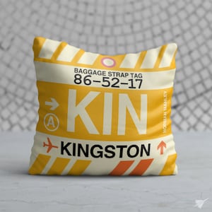 May include: A cushion with a luggage tag design. The cushion has a yellow background with white stripes and the text "KIN" and "KINGSTON" in black. The text "BAGGAGE STRAP TAG 86-52-17" is also visible.