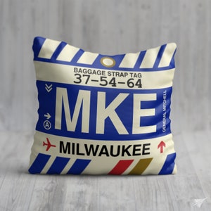 May include: A white pillow with a blue and white luggage tag design. The tag says "Baggage Strap Tag 37-54-64 MKE MILWAUKEE" and "General Mitchell" in smaller text.