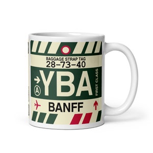 May include: White ceramic mug featuring a green and red luggage tag design. The tag reads "Baggage Strap Tag 28-73-40 YBA BANFF" with a red arrow pointing upwards and a small aeroplane icon.