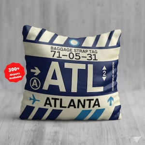 May include: A cream-colored throw pillow with a blue and white luggage tag design. The tag reads "Baggage Strap Tag 71-05-31 ATL Atlanta Hartsfield-Jackson".