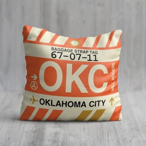 May include: A cream-coloured pillow with an orange and gold luggage tag design. The tag has the text "BAGGAGE STRAP TAG 67-07-11 OKC OKLAHOMA CITY" and "WILL ROGERS" on the side.