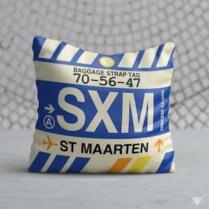 May include: A white and blue pillow with a luggage tag design. The tag says "SXM" and "ST MAARTEN" with the baggage strap tag number "70-56-47". The tag also says "Princess Juliana" and has an arrow pointing to the right.