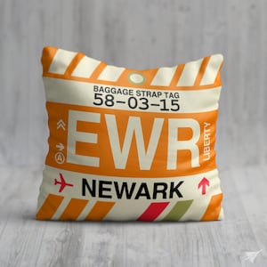 May include: Orange and white throw pillow with a luggage tag design. The tag says "EWR NEWARK" and "BAGGAGE STRAP TAG 58-03-15".