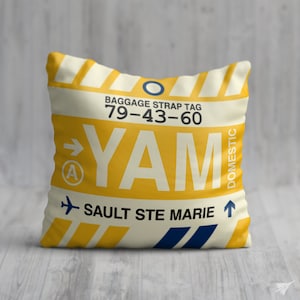 May include: Yellow and white cushion with a luggage tag design. The tag says "YAM" and "SAULT STE MARIE" with a baggage strap tag number "79-43-60".