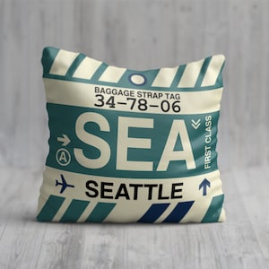 May include: A cream-coloured pillow with a blue and white striped design. The pillow features a luggage tag with the text "BAGGAGE STRAP TAG 34-78-06", an arrow pointing right with the letter "A" inside a circle, the text "SEA" in large white letters, an arrow pointing down with the text "FIRST CLASS", an airplane icon, and the text "SEATTLE" in black letters.