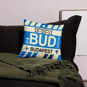 May include: A blue and white pillow with a luggage tag design. The tag says "BUD" and "BUDAPEST" with the airport code "10-22-11" and the airport name "FERENC LISZT".