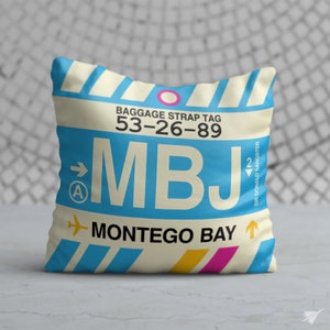 May include: Square cushion with a design resembling a baggage strap tag. The cushion features the text "MBJ MONTEGO BAY" in a blue and cream colour scheme, with a pink and yellow accent. The cushion also has the text "BAGGAGE STRAP TAG 53-26-89".