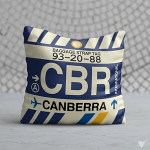 May include: A white pillow with a blue and yellow luggage tag design. The tag has the text "BAGGAGE STRAP TAG 93-20-88 CBR CANBERRA" and an arrow pointing to the right.