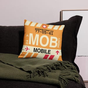 May include: Orange and white throw pillow with a luggage tag design. The tag says "BAGGAGE STRAP TAG 77-05-61 MOB MOBILE" with an airplane icon and an arrow pointing up.