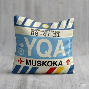 May include: A white and blue pillow with a luggage tag design. The tag has the text "Baggage Strap Tag 88-47-31 YQA MUSKOKA" and "LITTLE NORWAY" printed on it.