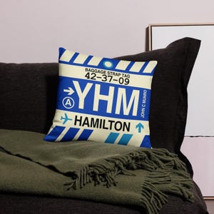 May include: A blue and white throw pillow with a luggage tag design. The tag says "Baggage Strap Tag 42-37-09 YHM HAMILTON" with an airplane icon and an arrow pointing right. The tag also says "John C Munro" in smaller text.