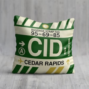 May include: A square cushion with a baggage tag design. The cushion displays the text "CID CEDAR RAPIDS EASTERN IOWA" in green and white, set against a cream background. The design incorporates a stylized airplane and arrow.