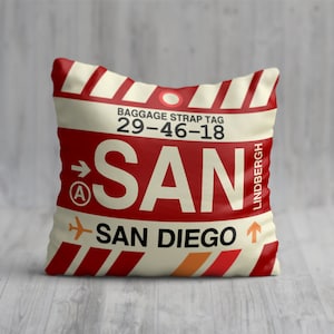 May include: A red and white striped throw pillow with a luggage tag design. The tag says "BAGGAGE STRAP TAG 29-46-18 SAN SAN DIEGO" with an arrow pointing to the right and a small airplane icon.