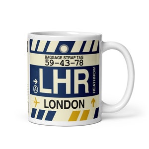 May include: White ceramic mug with a blue and yellow luggage tag design. The tag says "Baggage Strap Tag 59-43-78 LHR LONDON HEATHROW".
