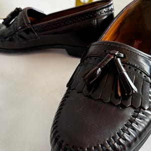 May include: A pair of dark brown leather loafers with tassel details. The shoes have a classic design with decorative stitching and a glossy finish. The loafers are displayed on a white surface.