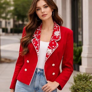 May include: A bright red blazer with white trim and gold buttons. The blazer's lapels feature a white and red polka dot pattern with floral accents. The blazer is worn over a white top and paired with blue jeans.