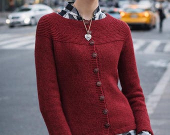 May include: A red knit cardigan with wooden buttons. The cardigan is worn over a black and white plaid shirt and dark jeans. A heart-shaped pendant hangs from a necklace. The background shows a city street.