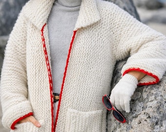 May include: A chunky off-white knit cardigan featuring a wide collar, two front pockets, and vibrant red trim along the edges. The garment is worn over a light gray turtleneck sweater and white pants, paired with white knit gloves and red sunglasses.