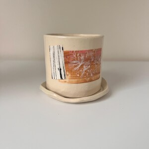 May include: A cream-colored ceramic cup with a matching saucer. The cup features an abstract design with black vertical lines and orange and white brushstrokes. The cup is cylindrical and sits on a small, oval saucer.