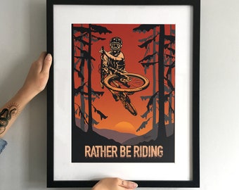 May include: A framed poster with a black frame and a white mount. The poster features a black and white illustration of a mountain biker in mid-air, with a sunset background and trees in the foreground. The text "Rather Be Riding" is printed in black at the bottom of the poster.