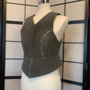 May include: A grey sleeveless vest with a V-neck and a fitted bodice. The vest is made of a thick fabric and has a textured surface. The vest is on a white mannequin.