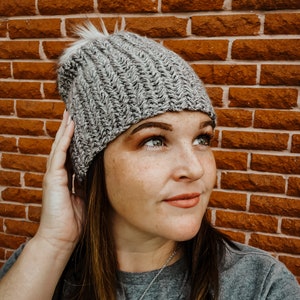 May include: A gray knit beanie with a white faux fur pom-pom. The hat has a ribbed design and is made of a soft, warm material. The beanie is shown against a brick wall.
