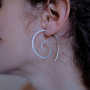 Puede incluir: Un pendiente de plata en forma de espiral con un diseño sencillo. El pendiente se lleva en la oreja izquierda.