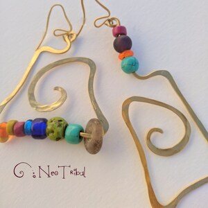 Puede incluir: Pendientes dorados de forma libre con cuentas de colores. Los pendientes presentan una textura martillada y un diseño abstracto único. Las cuentas son en tonos turquesa, morado, naranja, verde y azul. El texto "Neo Tribal" está en la esquina inferior izquierda.
