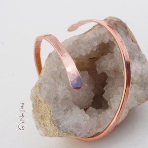 May include: Two copper bracelets, one with a hammered finish, are displayed on a white rock with a crystal-like texture.