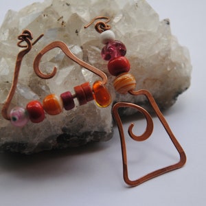 Puede incluir: Pendientes de alambre de cobre con un diseño geométrico y una cadena de cuentas de colores, incluyendo cuentas de color naranja, rojo, rosa y blanco.