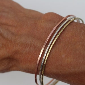 Könnte beinhalten: Drei dünne Metallarmreifen, die auf einem Handgelenk übereinander liegen. Die Armreifen sind aus Kupfer, Silber und Gold.