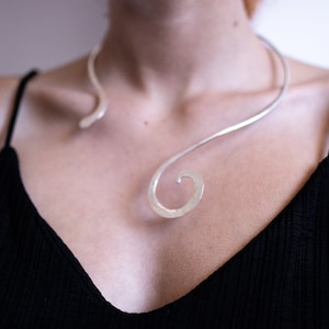 May include: A silver statement necklace with a unique, swirling design. The necklace is made of a thin, flexible metal and is worn around the neck.