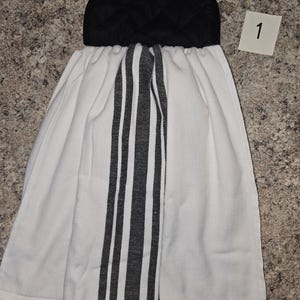 May include: A white kitchen towel with a black and white striped center panel and a black quilted top. The towel has a button closure and is designed to hang. A small white tag with the number "1" is in the upper right corner.