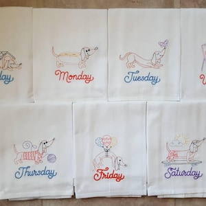 May include: Set of seven white tea towels with embroidered images of dachshunds and the days of the week. Each towel features a different dachshund activity, such as sleeping in a doghouse, eating a hotdog, painting, and skateboarding. The towels are embroidered with the words "Sunday", "Monday", "Tuesday", "Wednesday", "Thursday", "Friday", and "Saturday".