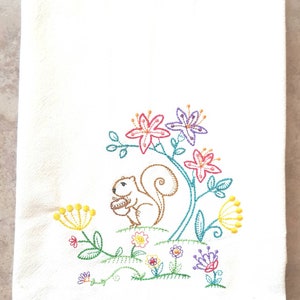 May include: Embroidered kitchen towel featuring a brown squirrel holding a nut, surrounded by colourful flowers in shades of pink, purple, yellow, and green.