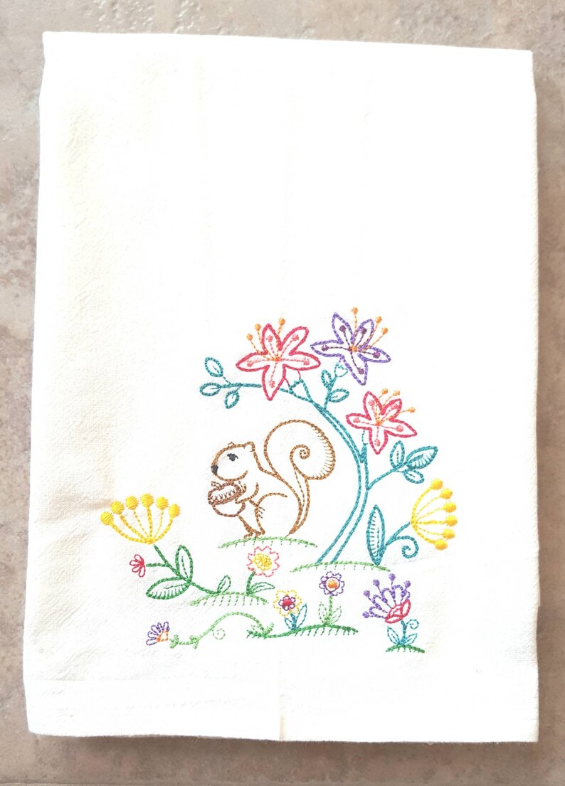 May include: Embroidered kitchen towel featuring a brown squirrel holding a nut, surrounded by colourful flowers in shades of pink, purple, yellow, and green.