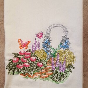 May include: White kitchen towel with an embroidered design of a garden scene. The design features a stone archway with flowers and butterflies, a path, and a variety of plants in shades of green, pink, purple, orange, and blue.