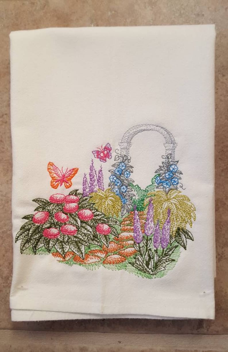 May include: White kitchen towel with an embroidered design of a garden scene. The design features a stone archway with flowers and butterflies, a path, and a variety of plants in shades of green, pink, purple, orange, and blue.