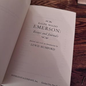 May include: Open book with the title "EMERSON: Essays and Journals" by Ralph Waldo Emerson. The book is published by Doubleday & Company, Inc. The text is in black font on a cream-colored page. The book is open on a wooden surface.