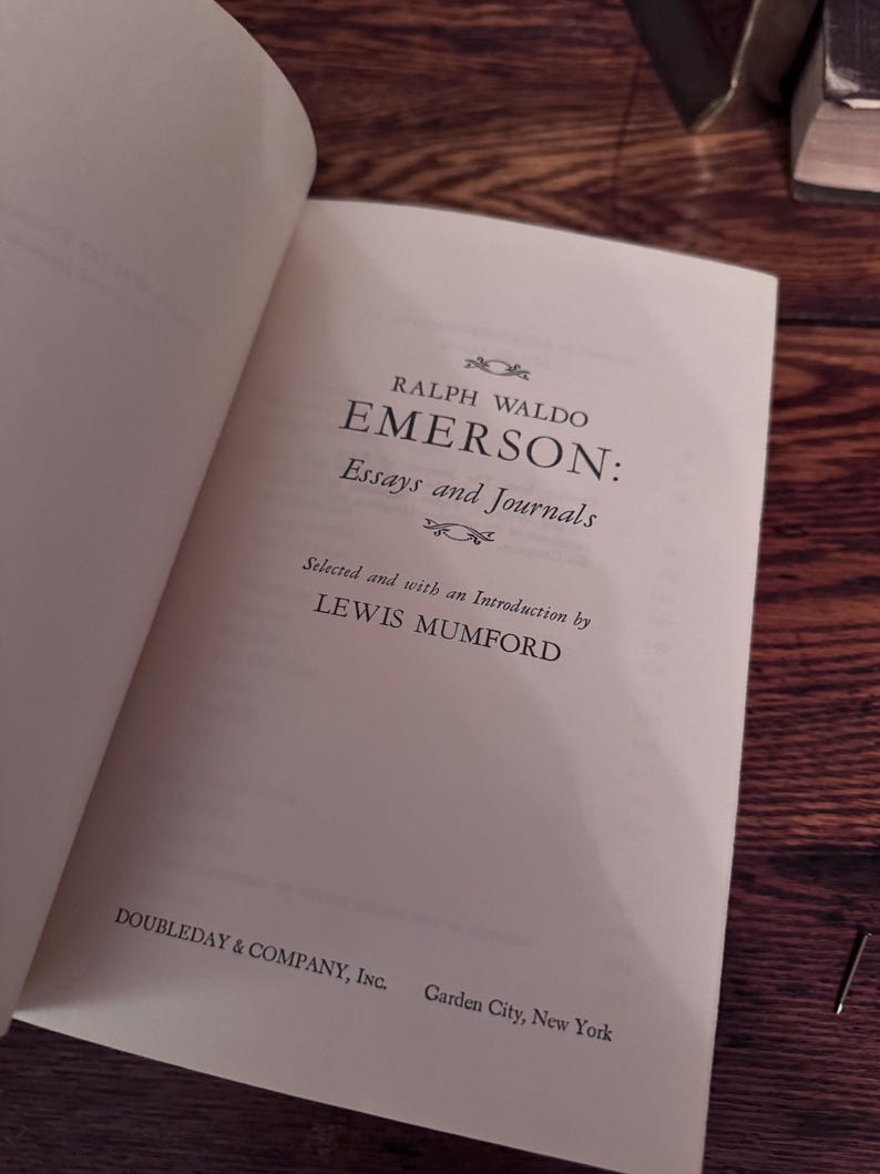 May include: Open book with the title "EMERSON: Essays and Journals" by Ralph Waldo Emerson. The book is published by Doubleday & Company, Inc. The text is in black font on a cream-colored page. The book is open on a wooden surface.