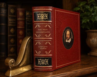 May include: A red leather-bound edition of "The Yale Shakespeare: Complete Works" with gold lettering and accents. The book features a portrait of Shakespeare and is propped up by a gold-colored bookend. Other books are visible in the background.